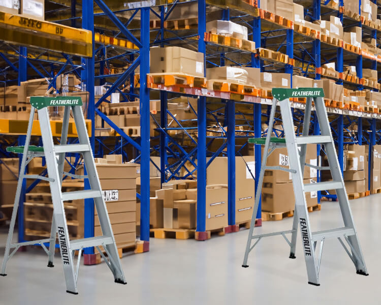 Featherlite step ladders set up inside a large warehouse with tall shelving units - Durable Featherlite industrial ladders