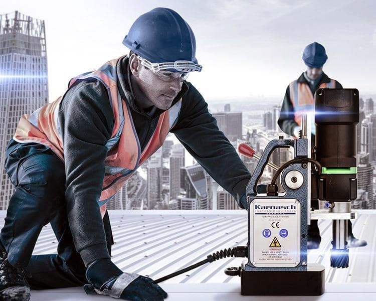 Construction worker in helmet and reflective vest operating a Karnasch mag drill on a rooftop with a cityscape in the background, emphasizing safety and precision tools