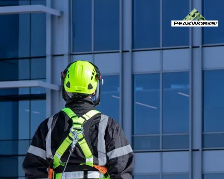 Worker in Peakworks safety gear, including a helmet and harness, inspecting a large glass building with the Peakworks logo visible, emphasizing industrial safety