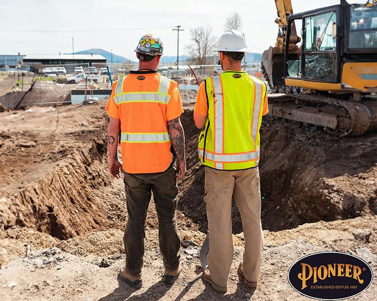 Two construction workers in Pioneer high visibility vests and hard hats near an excavation site, with a bulldozer in the background, discussing the project