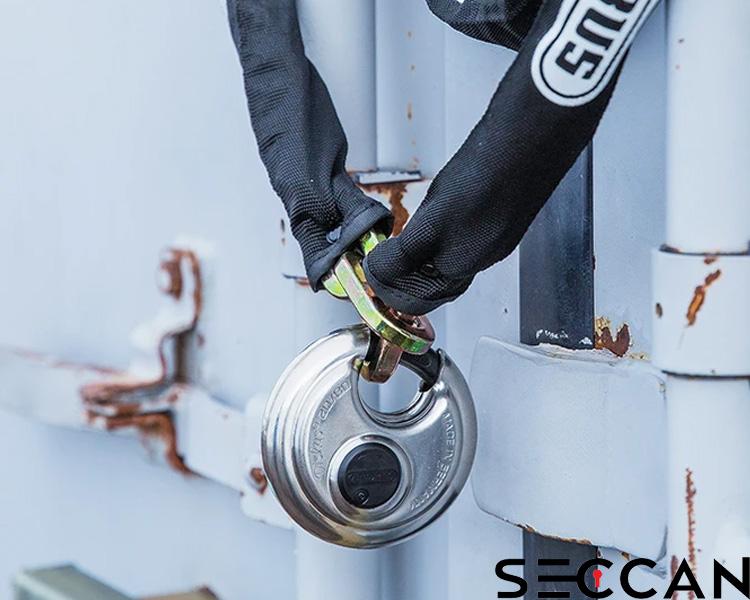 Person wearing black gloves locking a silver Seccan padlock on a rusted metal gate, showcasing Seccan branding.